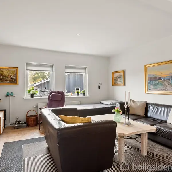 En stue med sort lædersofa, placeret omkring et lyst sofabord. Væggene har malerier, og vinduerne giver udsyn til en have. TV og reoler supplerer indretningen.