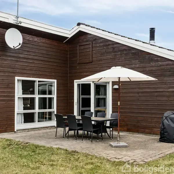 Træhus med betonterrasse har parasol og seks sorte stole omkring bordet; grill dækket til med et omslag. Græsplæne omgiver området, og en parabolantenne ses på taget.