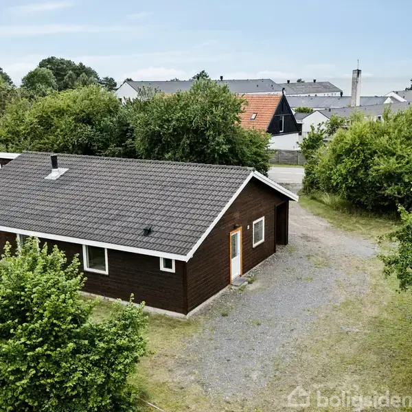 Et mørkt træhus står stille omgivet af frodige buske og træer. En grussti fører til døren, mens flere huse ses i baggrunden.