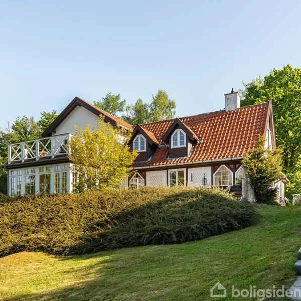 Et hvidt hus med rødt tegltag står på en skrånende græsplæne. Det har en balkon og er omgivet af buske og træer under en klar blå himmel.