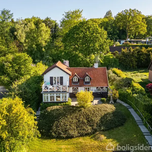 Huset med rødt tag og hvide vinduer står omgivet af grønne træer og buske. Stenstien fører til indgangen i den frodige have.