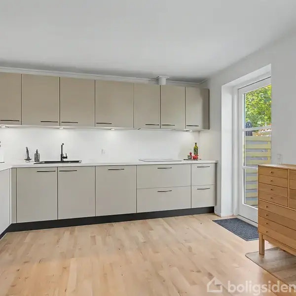 Et moderne køkken med lyse skabe og indbygget ovn på venstre side. En kommode med vase og blomster står ved siden af en glasdør ud til en have.