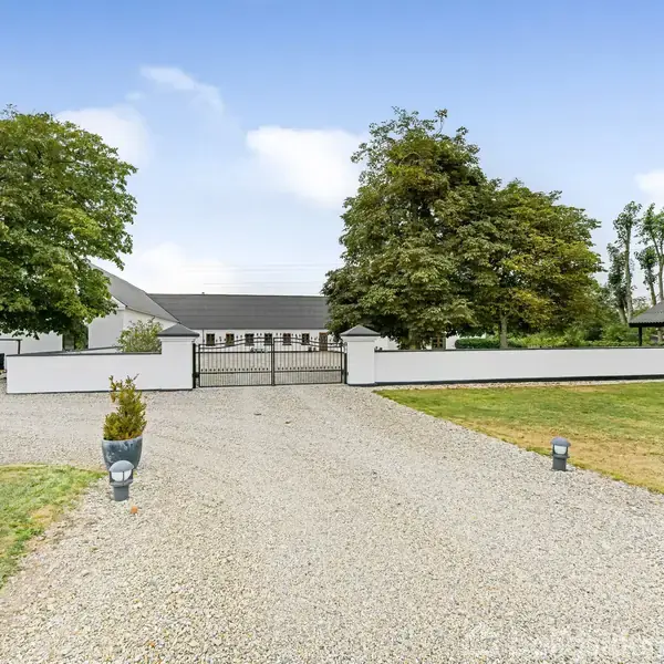 Gårdsplads med grusvej fører til hvidbygning mellem store træer; grøn plæne ligger ved siden af. Bygningen ligner en traditionel gård eller landhus med port indgang.