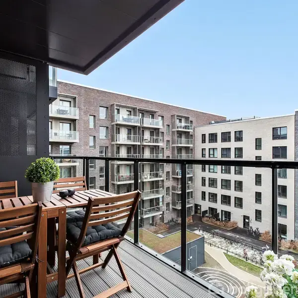 Træmøbler står på en balkon med glasrækværk. I baggrunden ses moderne boligblokke på en klar dag. En lille grøn plante står på bordet.