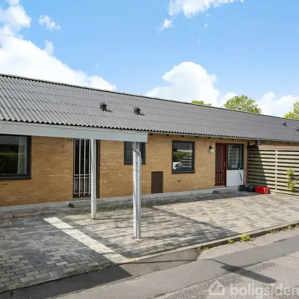 En rækkehusbygning med lyse mursten og sort tag, står stille langs en vej. Foran er der en parkeringsplads med fliser. Der er klar blå himmel med skyer i baggrunden.