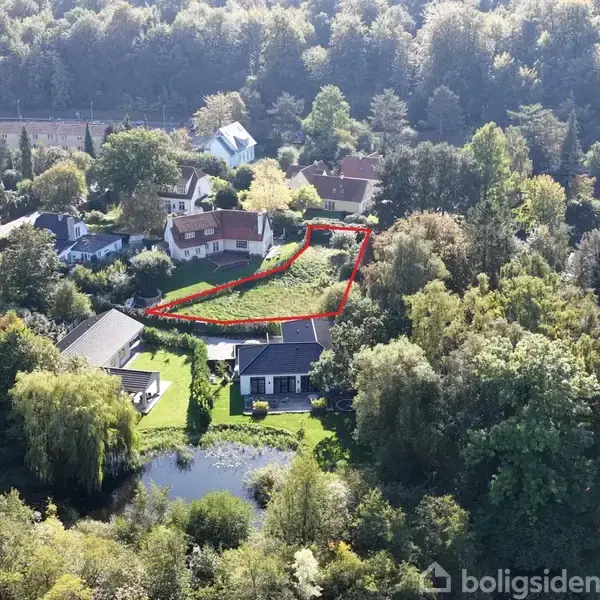 Grunden markeret med en rød streg ligger i et grønt, skovrigt område med forskellige huse omkring. En nærliggende dam er synlig blandt træerne.