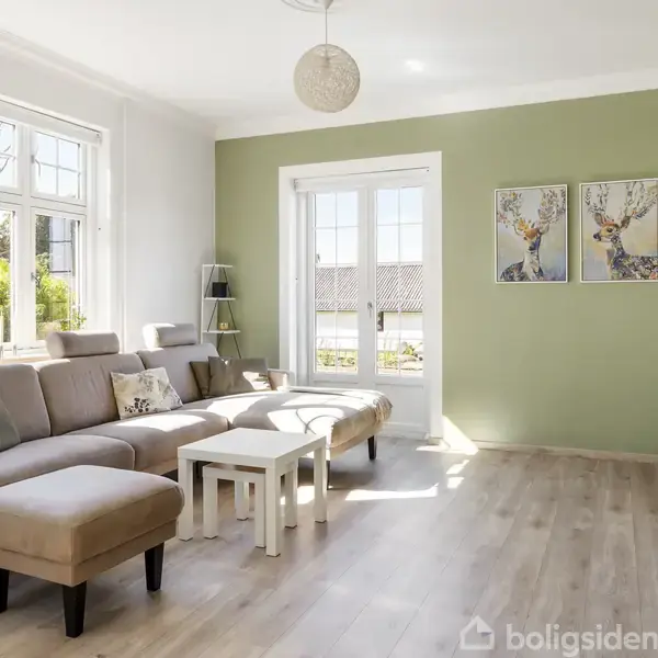En beige sofa står i en lys stue, der åbner op mod en grøn væg med to hjortemalerier. Store vinduer og en glasdør giver udsigt til en terrasse.