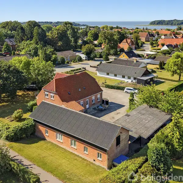 Hus med rødt tegltag står mellem grønne haver i et boligkvarter. Flere huse er synlige i nærheden, og der er udsigt til en skov og et vandbassin i baggrunden.