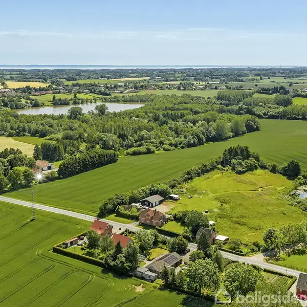 Landsby med spredte huse omgivet af grønne marker og skove. En vej snor sig gennem området, og der er en sø i baggrunden under en blå himmel.