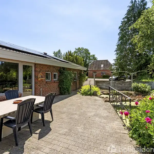 Terrasse med borde og stole ligger foran et murstenshus med store vinduer. Omkring er en have med blomster og store træer, der skaber en rolig og grøn atmosfære.