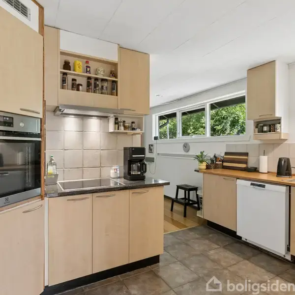 Køkken med lyse træskabe og sorte bordplader. En ovn og en mikrobølgeovn er indbygget. På væggen er hvide fliser. Et stort vindue i baggrunden lyser rummet op.