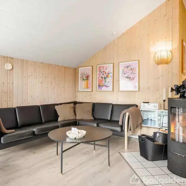 En sort sofa sidder i et hjørne med farverige billedrammer over sig; en brændeovn brænder på en flisebelagt overflade ved siden af, i et lyst træpanelrum med store vinduer.