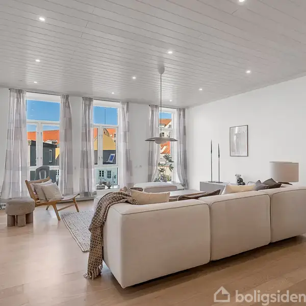 En lys stue med en stor, hvid sofa centreret mod et sofabord og en stol nær vinduerne. Loftslamper oplyser det moderne, stilfulde rum med trægulv.