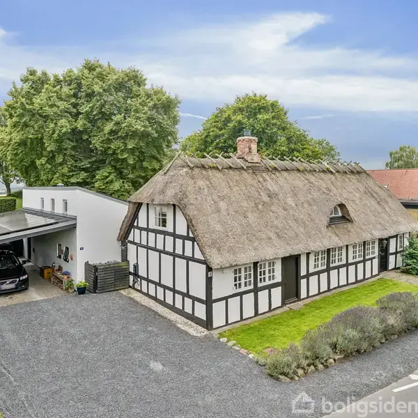 Bindingsværkshus med stråtag står stille ved siden af en moderne garage med bil, omgivet af grusvej og grøn beplantning; baggrunden har træer og flere huse.
