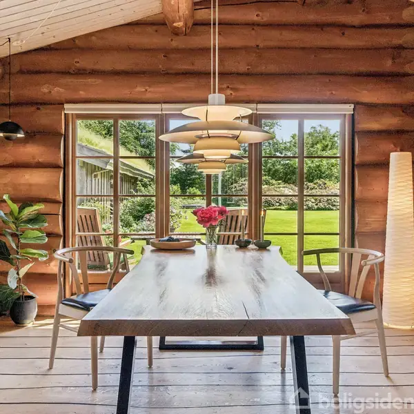 Træbord er dækket med blomster, omgivet af stole. Det står mellem vinduer med udsigt til en grøn have. Trævægge og loft med en hængende lampe skaber en rustik atmosfære.