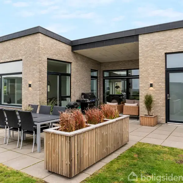 Moderne hus står stille på en flisebelagt terrasse med spisebord og planter i kasser; omgivet af en velplejet græsplæne under en klar blå himmel.