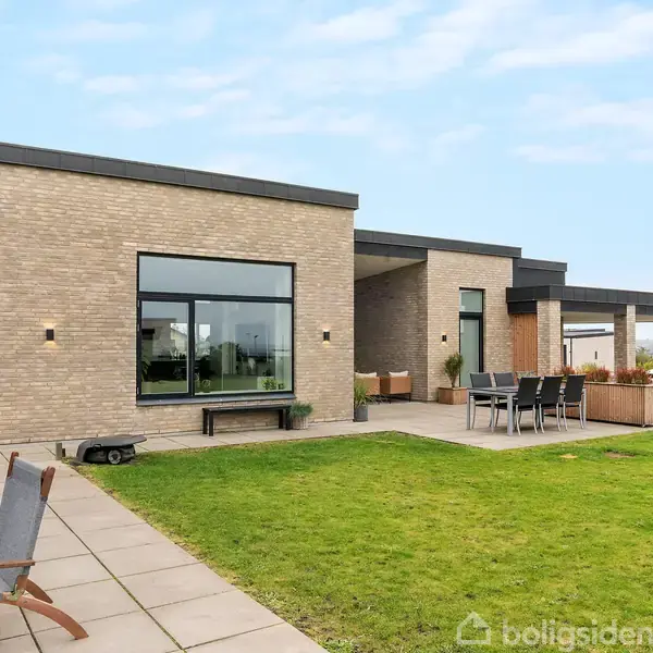 En moderne murstensvilla står på en velplejet græsplæne, omgivet af en stenbelagt terrasse. Et bord med stole står udenfor, mens loungestole er placeret nær huset.