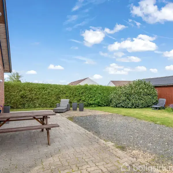 En træbænk står på en flisebelagt terrasse ved siden af et rødt murstenshus. I baggrunden ses en grøn hæk, en græsplæne og to udhuse under en blå og skyet himmel.