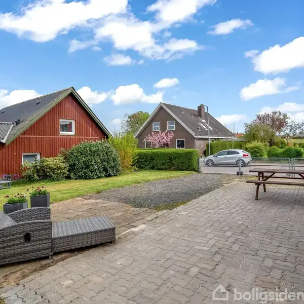 To liggestole står på en terrasse ved siden af et rødt hus. I baggrunden ses en bil på en grusvej og flere huse med haver og buske.