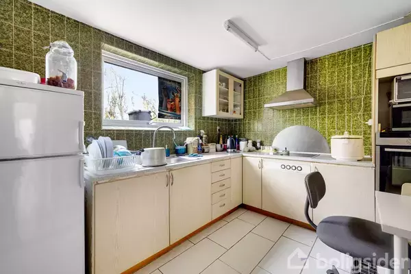 Køkken med hvide skabe og grønlige fliser, besidder et gaskomfur og en emhætte. Ved siden af står en høj køleskabsfryser kombination. Et vindue lader lys komme ind i rummet.