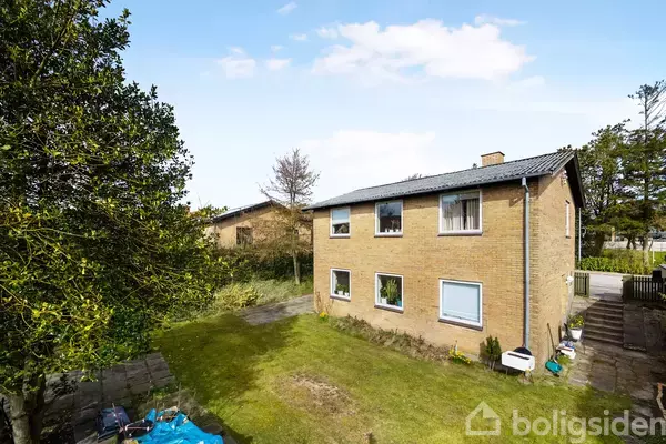 Et to-etagers murstenshus står i en solrig have med grønne planter og en terrasse. Der er græsplæne omkring huset, og træer omkranser området.