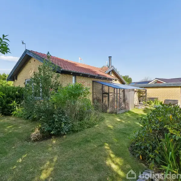 Gult murstenshus med rød tag, omgivet af grøn have og buske. Drivhus ved siden af huset. Klart solrigt vejr og blå himmel.