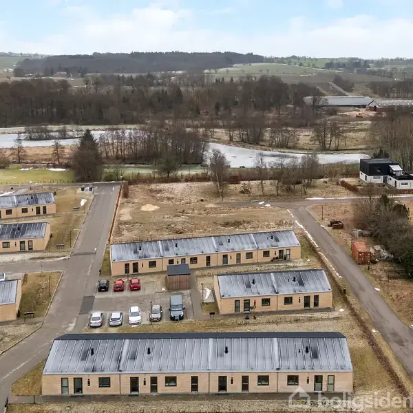 Flere rækkehuse står i rækker med vejforbindelser, omgivet af en landlig scene med en sø og skov i baggrunden. Biler er parkeret foran husene.