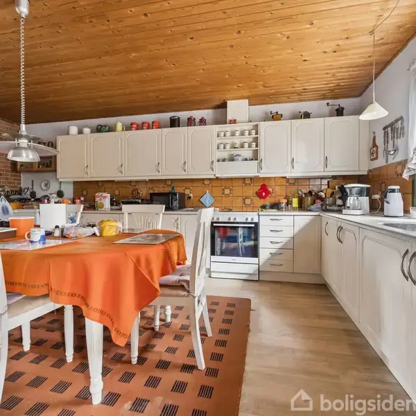 Et køkken med et spisebord dækket med en orange dug. Bordet er omgivet af hvide stole. Der er hvide skabe, en ovn og en kogeplade. Vinduet har gardiner.
