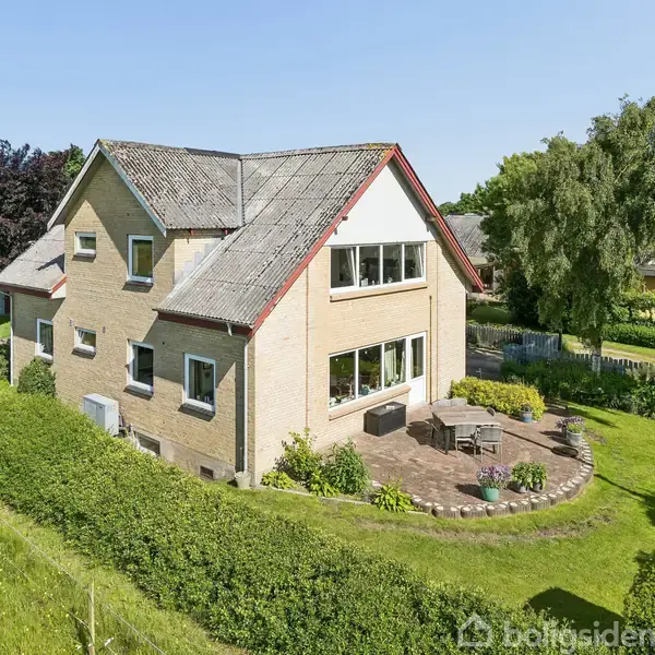 Hus med skråt tag, omgivet af grøn have og buske, placeret i et landområde. Et terrasseområde foran huset har havemøbler.