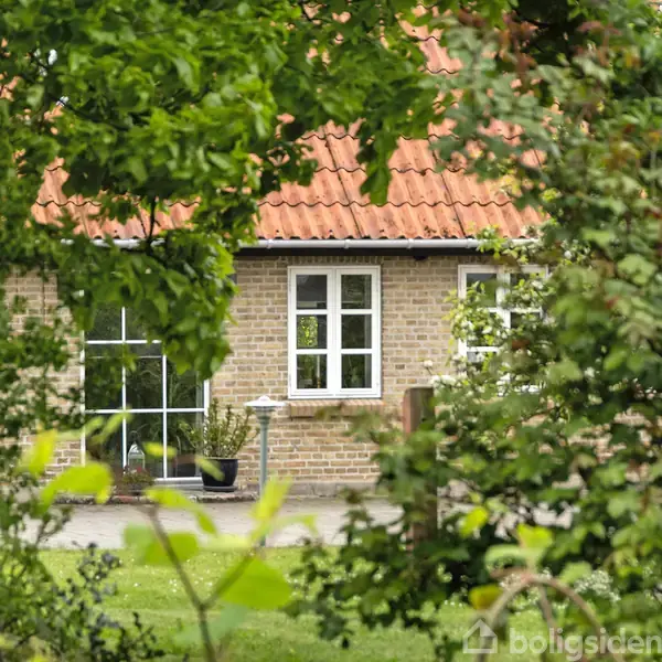 Et murstenshus med hvide vinduer står i en frodig have. Røde teglsten dækker taget, omgivet af tætte, grønne træer og buske. Græsset er velplejet omkring huset.