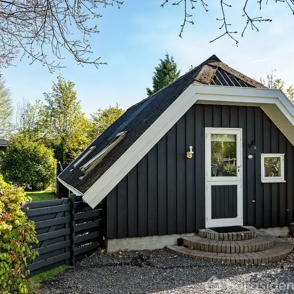 Et lille sort, trekantet hus med skråtag står i en grøn have. Huset har en glasdør og et lille vindue. Grussti fører til indgangen.