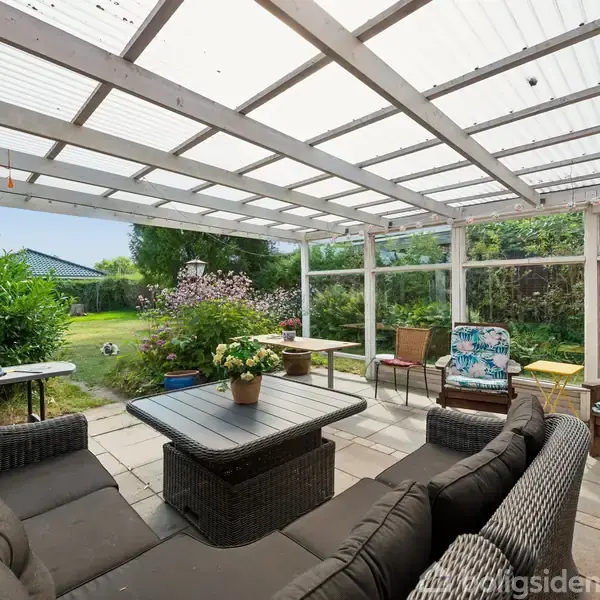 Overdækket terrasse med kurvemøbler omkring et lavt bord med blomsterpotte. Møblerne vender mod en grøn have med græsplæne og buske. En grill står til venstre.