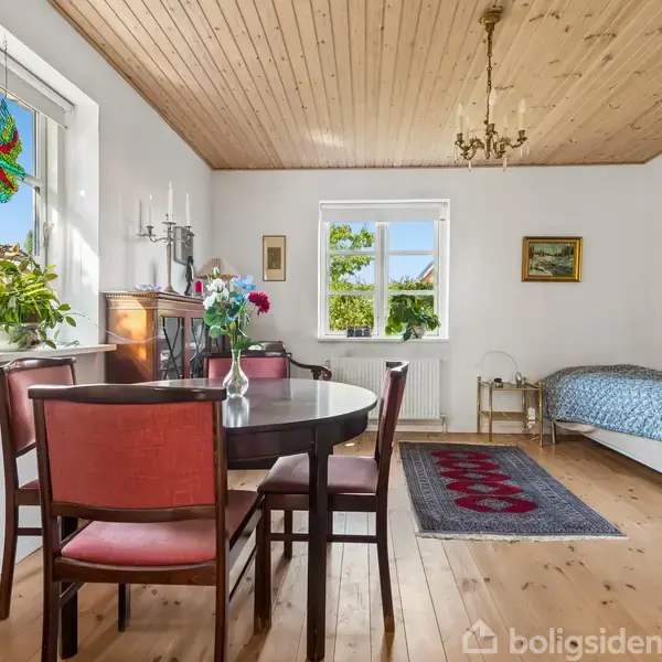 Spisebord med vase står i et lyst rum med træloft. Ved siden af er en enkeltseng, og vinduer med udsigt. Et tæppe ligger på trægulvet, og planter pryder vindueskarmen.
