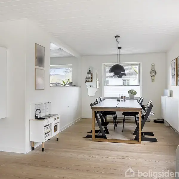 Spisestue med træbord omgivet af sorte stole. Hængelampe over bordet. Lysindfald fra store vinduer. Hvidt kabinet og dekorationer i hjørnet. Blomster i en blå vase på vindueskarmen.