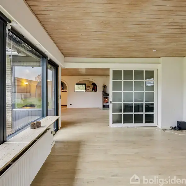 En lys stue har store vinduer til venstre og en skydedør i glas mod andre rum. Til højre står en kontorstol foran et skrivebord med en computerskærm.