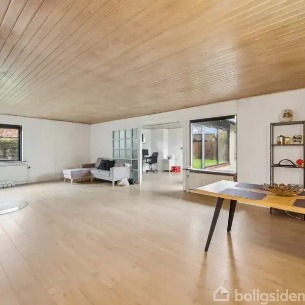 Rummeligt værelse med træloft og lyst trægulv indrettet med enkelt bord og stol. Store vinduer giver udsigt til en grøn have. En sofa står ved væggen, og der er en reol.