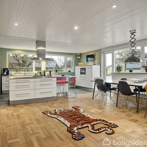 Køkken med en central ø omgivet af lyse møbler, planter og en tigerformet tæppe på trægulvet. Store vinduer giver udsigt til haven.