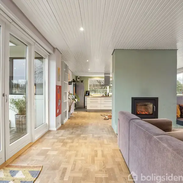 Stue med grå sofa og pejs på grøn væg, overfor store vinduer med udsigt til terrasse. Køkken i baggrunden. Planter hænger fra loftet, og farverige puder pryder sofaen.