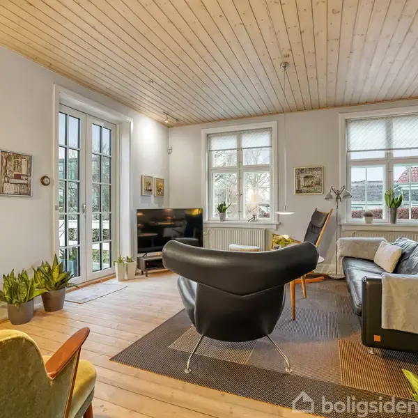 En moderne stue møbleret med sorte og grå sofaer og en lænestol i midten. Store vinduer og planter giver lys og liv til rummet. Trævægge og gulv skaber en hyggelig atmosfære.