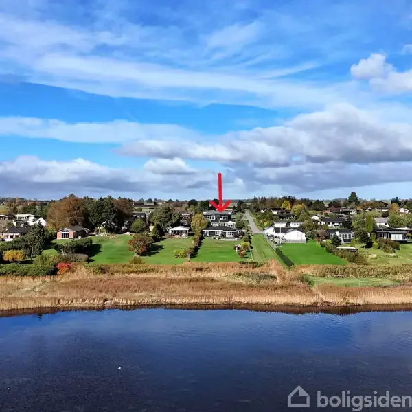 En rød pil peger mod et hus i et naturskønt område, omgivet af grønne træer og græs. Foran ses en bred flod under en blå himmel med spredte skyer.