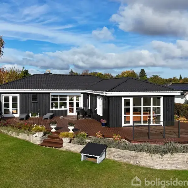 Sort træhus med store vinduer står på en hævet terrasse omgivet af en frodig have. Himlen er blå med spredte skyer. En mindre bygning ses på græsplænen.