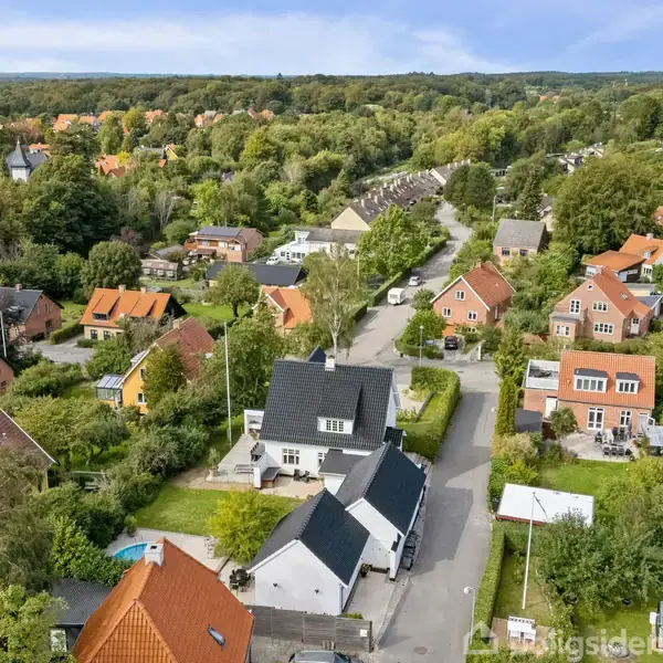 Huse står stille i et grønt boligkvarter omgivet af træer og veje. Små haver omkranser husene, og en skov ses i horisonten.
