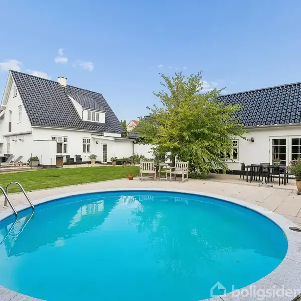 Rund swimmingpool med stige er omgivet af terrassefliser. I baggrunden ses en hvid villa med sorte tage og en have med græs og træer. Stole og borde står ved siden af.