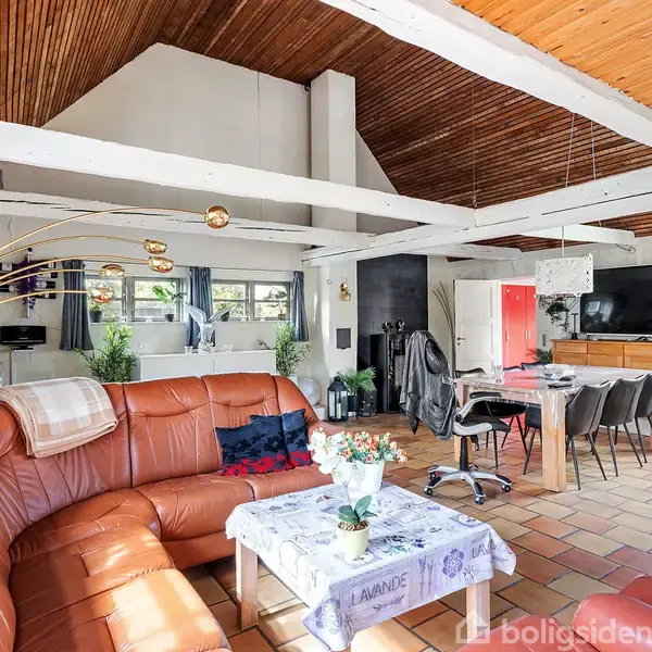 En stor hjørnesofa i brun læder dominerer stuen, hvor et sofabord står foran. Ved siden af er et spisebord med stole. Rummets loft er med træpaneler og hvide bjælker.