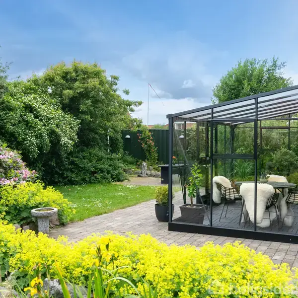 Et drivhus af glas står på en flisebelagt terrasse. Indeni er der møbleret med et bord og stole. Omgivet af frodig have med blomster og buske under en klar, blå himmel.
