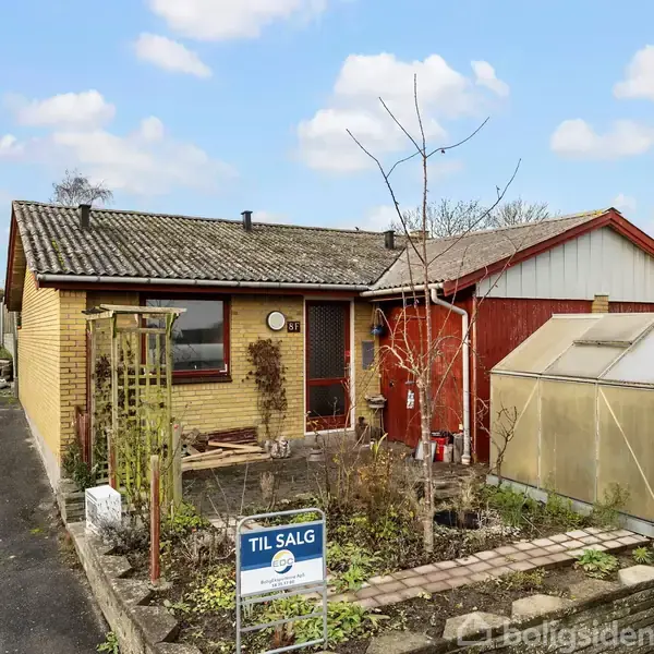 Huset har en gul murstensfacade og saddeltag, ved siden af et drivhus. Foran er et "Til salg" skilt fra Realmæglerne. En rød bil er parkeret til venstre.