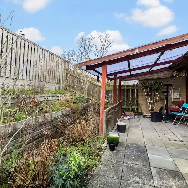 En overdækket terrasse med borde og stole ligger ved siden af et hus. Haven har stentrapper og blomsterbede. Et hegn omgiver området i en solrig atmosfære.