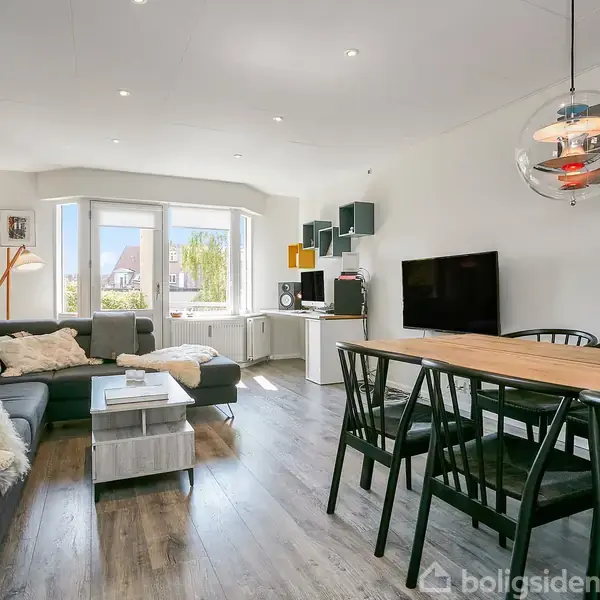 En moderne stue med et L-formet gråt sofaarrangement, trægulv, og stort vindue. Rundt bord ved sofaen, spisebord med sorte stole, fladskærms-TV og dekorative lamper i loftet.