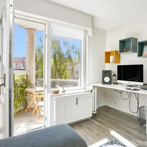 En moderne stue med en computer og højttalere på en hvid skrivebord, åbner ud mod en balkon med et lille cafébord og stole. Sollys skinner gennem det store vindue.