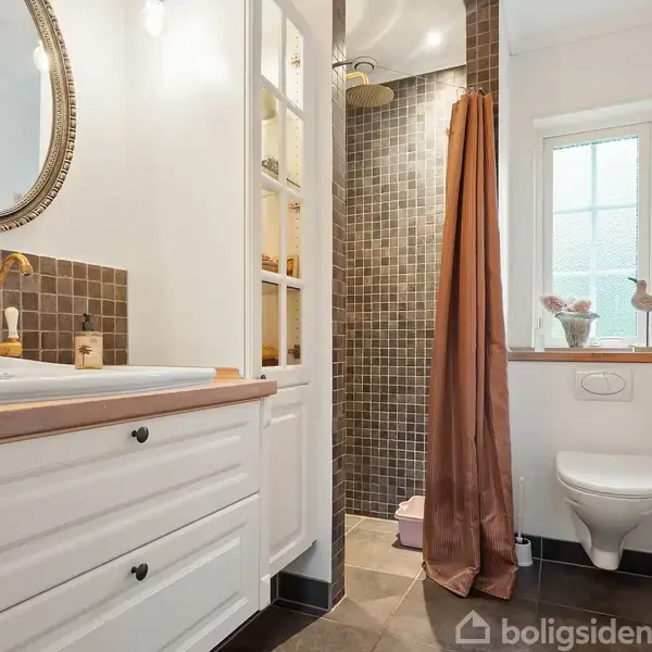 Et badeværelse med en hvid vask og gyldent armatur anbragt foran et spejl. Brusekabine med brunlige fliser. Ved siden af hænger et lysebrunt forhæng, og et vindue med lys indfald giver naturligt lys.